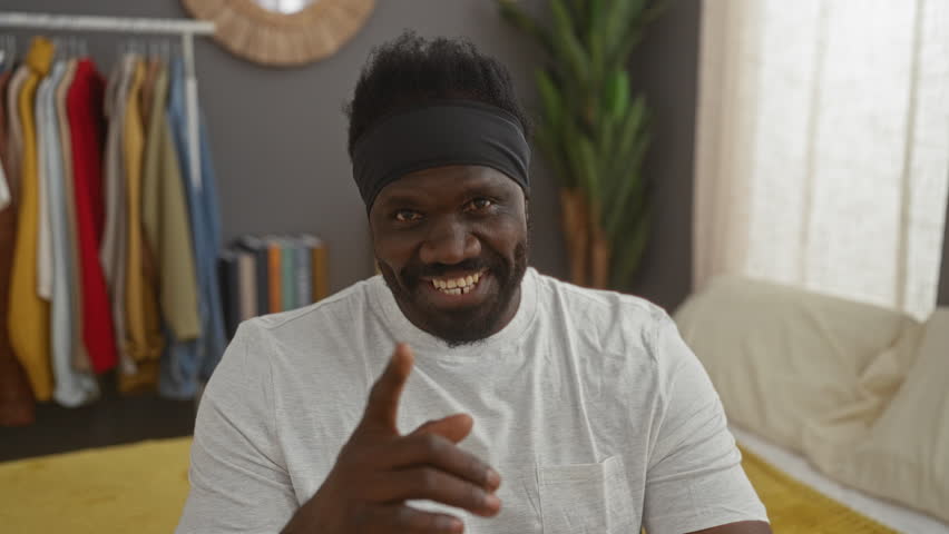 Handsome, young african american man in white shirt smiling and waving while sitting on a bed in a cozy, stylish bedroom.