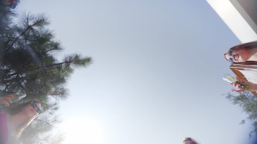 Women friends clink glasses with champagne and drink for good, fun summer holidays in country luxury club. Models in bright bikinis relaxing with alcohol and waving in camera. Shoot from below.