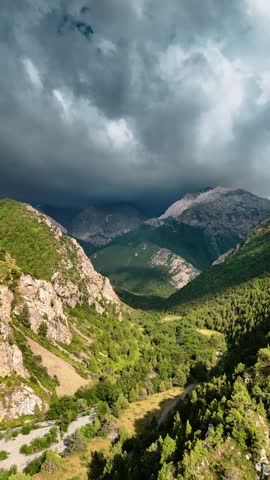 Mountains covered with coniferous trees rise to the cloudy sky, creating a picturesque green landscape. Clouds swirl over the peaks, giving the scene drama and mystery.