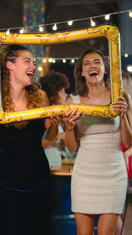 Vertical video of female friends having fun posing with photo booth photo frame at party in bar - shot in slow motion