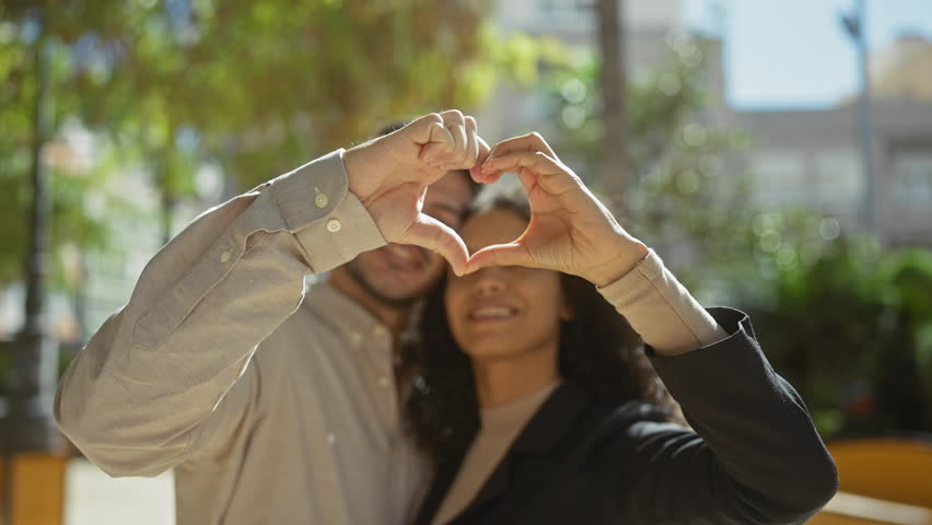 A loving couple forms a heart shape with their hands while enjoying a sunny day outdoors in a lush, green park.