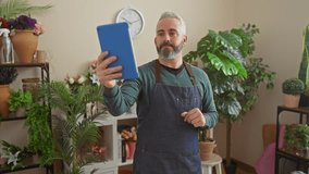 A bearded man holds a tablet for a video call in a lush flower shop with various plants in the background. - Powered by Shutterstock - Get 15% off with code: PIKWIZARD15