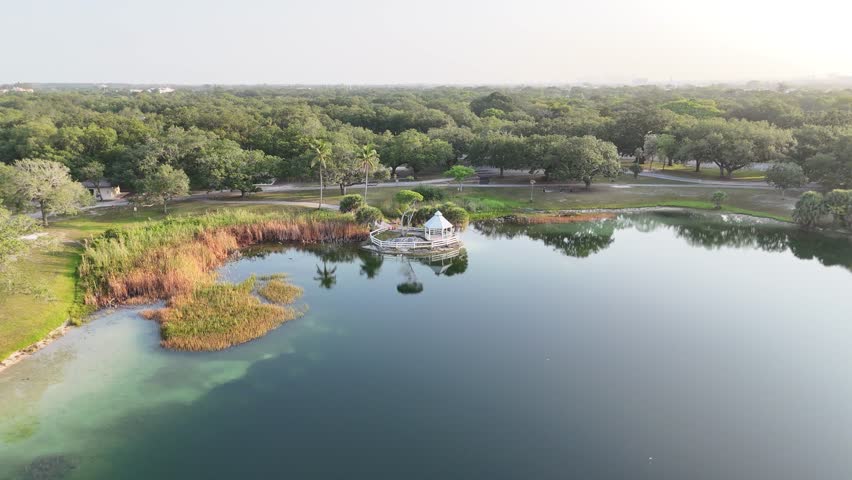 Aerial drone view of T.Y. (Topeekee Yugnee) Park in Hollywood, Florida, with a serene lake, gazebo, and The Guitar Hotel in the distance.