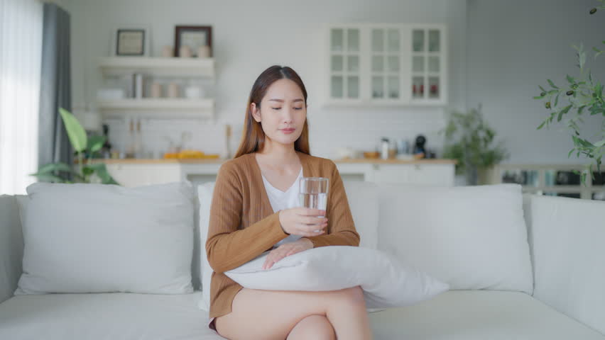Young Asian woman smiling while sitting on sofa happy expression, hand holding water glass and drinking water