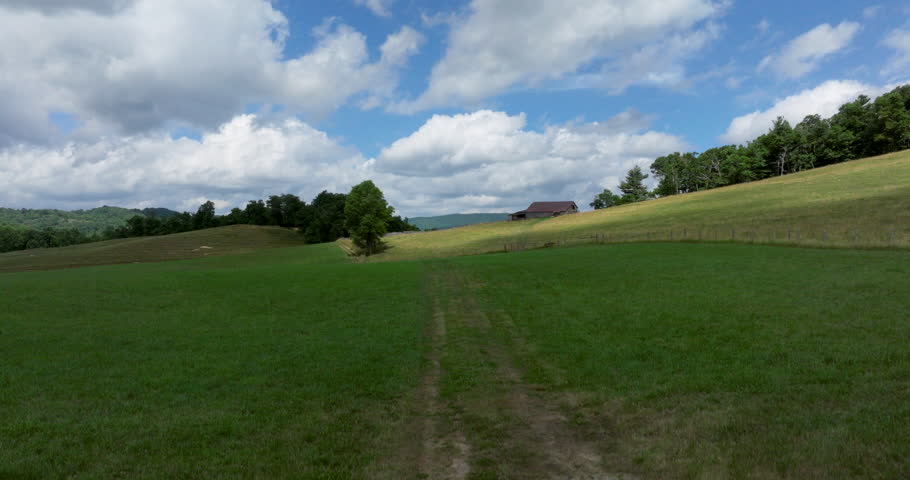 A cinematic drone ascends in a mountain landscape on a Summer day with an old barn in a pasture and a mountain range in the distances as clouds pass by in this peaceful scene and epic view of nature.