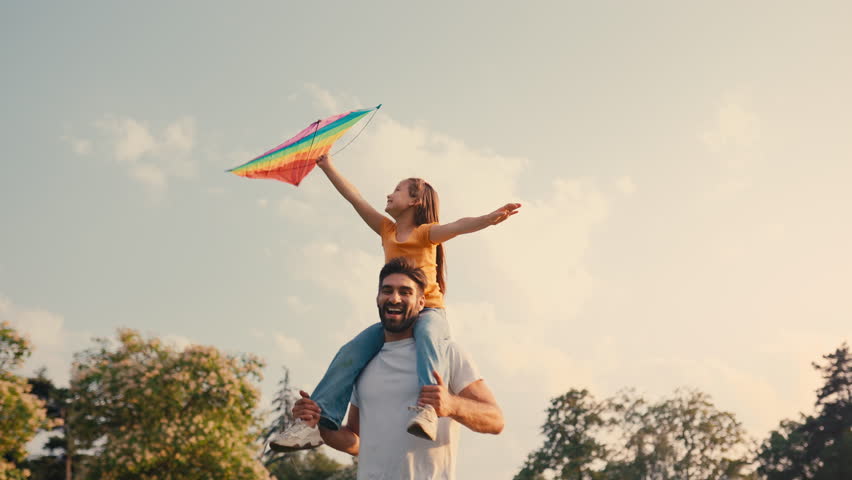 At the park, a father carries his daughter on his shoulders as she flies a kite, creating cherished outdoor memories. Their bonding fosters a strong relationship