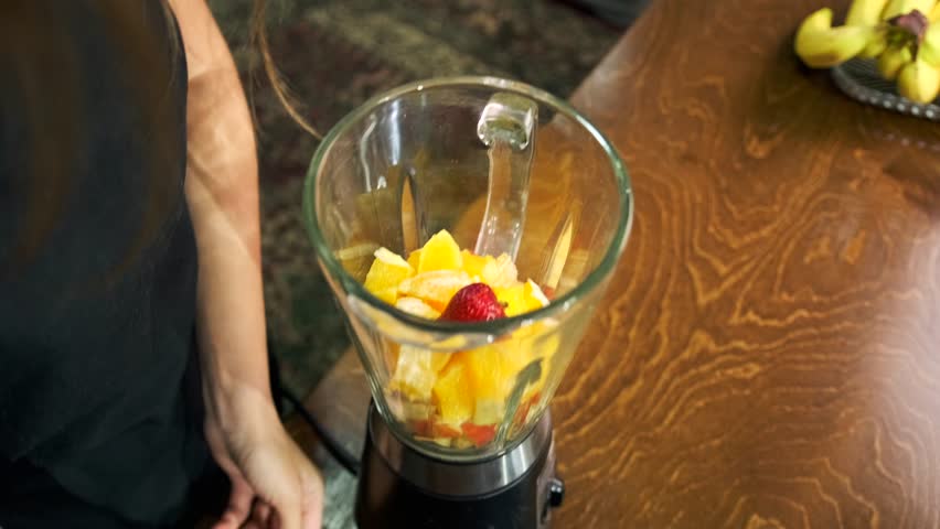 Peeling strawberries with the hands of a young woman to prepare a smoothie with orange in a blender lying on a wooden kitchen counter close-up.