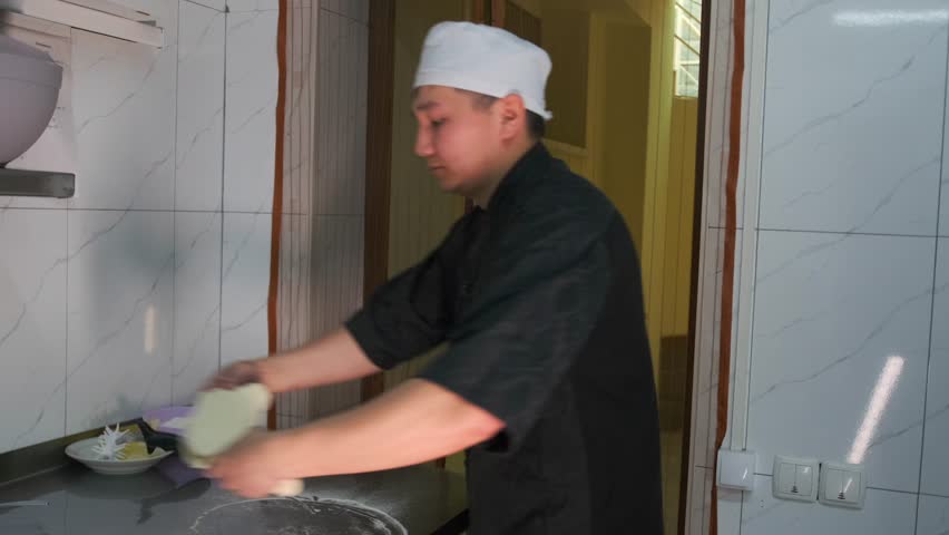 Man chef preparing pizza dough on a restaurant kitchen counter next to a container with ingredients. Side view.