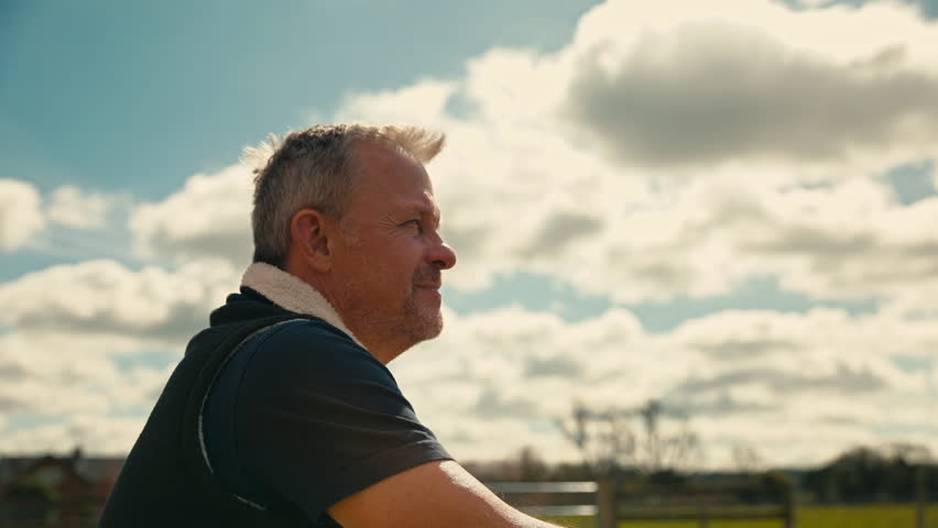 Mature male farm worker standing outside barn leaning on fence looking over fields - shot in slow motion