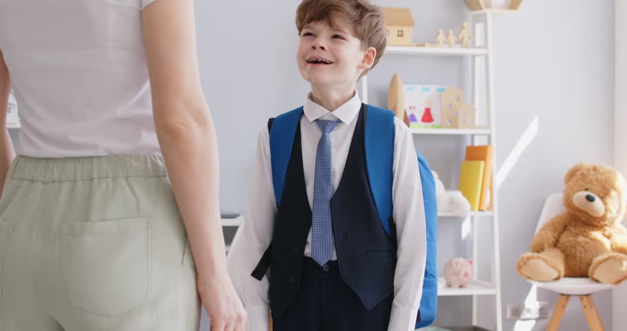 Positive mother preparing young schoolboy son, student or pupil with his backpack for school, hugging him on at home. Tender moment of a mother care, goodbye and support before child day at school.