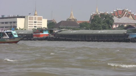 Barges and tourist boats pass on the Chaopraya River in Bangkok with the Grand Palace is in the background.