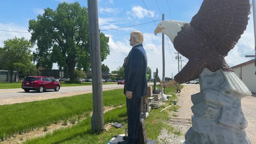 Iowa - 6.24.2024 - cars drive past an iowa roadside shrine to donald trump, which includes a statue of him and a bald eagle.