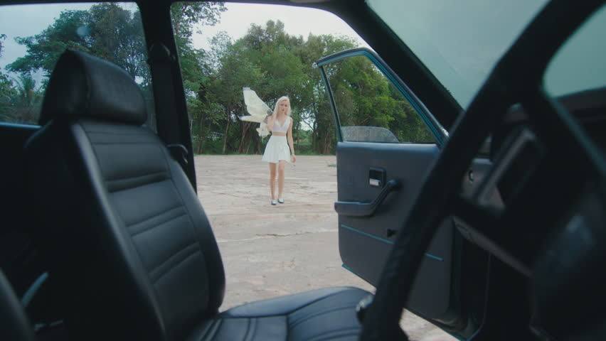 A woman wearing a white dress is strolling towards an open car door in a tranquil rural setting