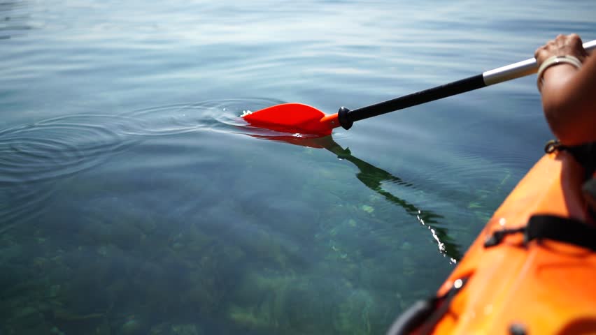 A person is paddling a kayak in the water. The water is calm and the sun is shining brightly. The person is wearing a black life jacket and a white wristband.