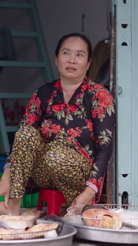 Barefoot mature Asian woman seated next to a fan waits for customers at a stall in the local market on Ho Chi Minh City street in Vietnam