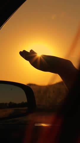 Woman driver sun. Free young woman travels by car, extends her hand to sun from car window. Vacation. Young girl with sits in front seat of car, her hand out window, catching glare of setting sun.