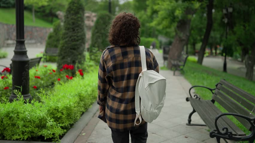 Back view A brunette girl in a checkered shirt with a white backpack walks through the park after classes at the university