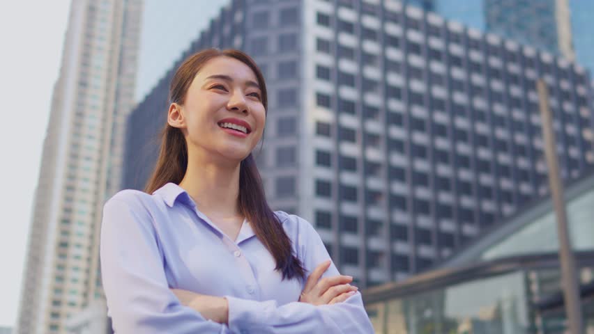 Asian youg beautiful businesswoman standing outdoor in city skyscrapers. Attractive employee female worker feeling happy and confident then smile, crossing arms with confidence at high rise buildings.
