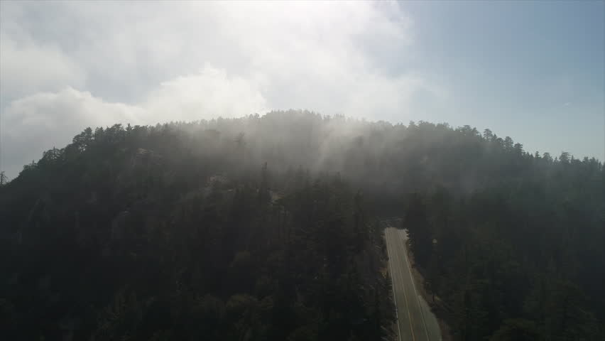 Slow tilting up drone shot of fog rolling over a mountain top and an empty road. Shot in the Angeles National Forest.