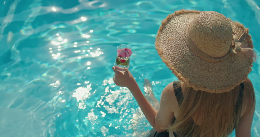  Young woman in a hat holding a cool drink, sitting on the edge of a pool on a hot summer day