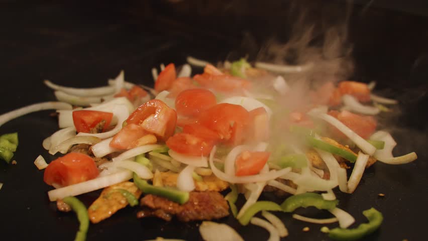 A slow-motion closeup of mushrooms falling on Chicken Fajita vegetables and meat being prepared on grill with smoke coming out of it