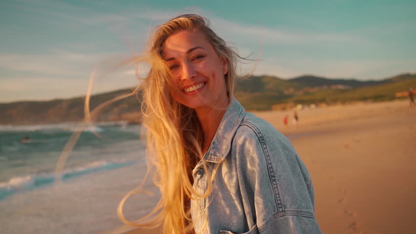 Woman smiling looking camera standing on ocean shore, wind blowing her hair. Portrait shooting happy blonde woman on beach. Young girl in denim shirt enjoying ocean views during sunset.