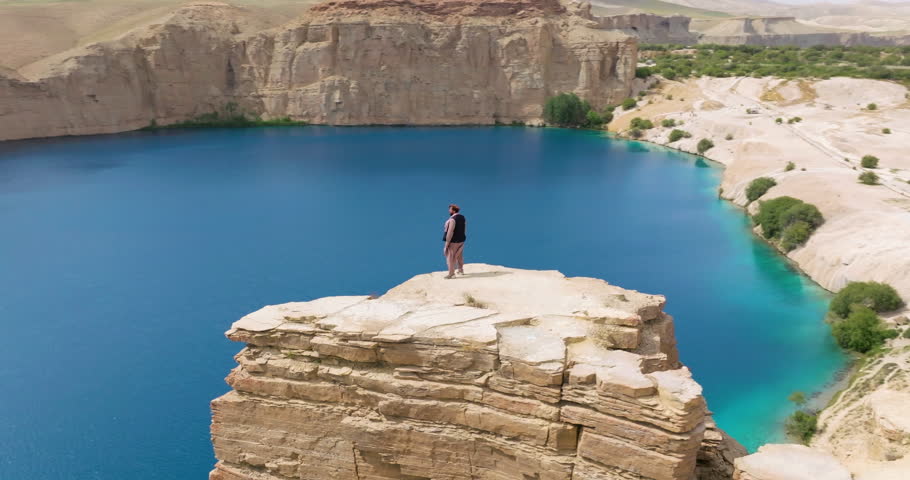 Male Hiker Stands On Clifftop With Scenic Lake View At Band-e Amir National Park In Afghanistan. aerial orbiting shot