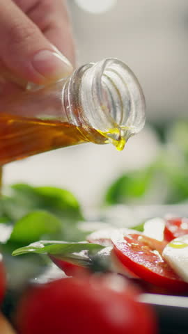 Vertical position. A housewife adds olive oil to a salad on the kitchen table. A young woman pours olive oil using various vegetables and cheeses to create a mozzarella salad