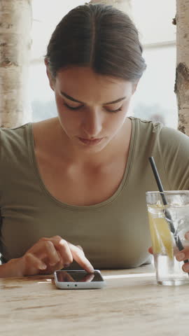 Young beautiful woman sitting in a cafe, smiling and using her smartphone to browse the internet, text, chat with friends, or check social media news. Tapping on the screen and Drink a glass of water.