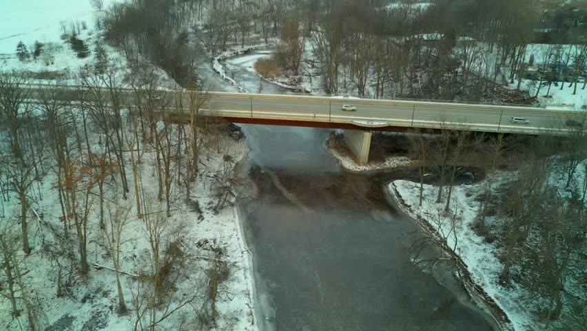 An aerial view flying up to a person on a bridge as they look down at the frozen river. The person looks over the edge and then continues to run