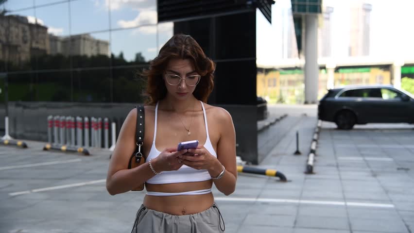 Portrait of beautiful long haired brunette woman in eyeglasses and hip hop streetwear standing by modern glass buildings and holding her smartphone and handbag on city street. Soft focus.