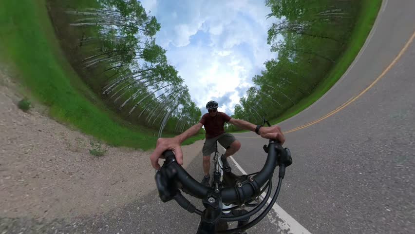 Extreme wide-angle view from a 360 degree camera looking at a male cycler pedalling along the side of a paved road that runs through a green forest.  

