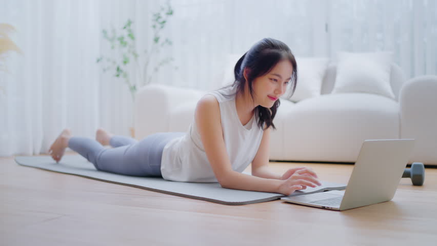 Young Asian woman lying on the floor on yoga mat in front of laptop and training at home. Beautiful smiling woman doing stretching exercise while communicates with a trainer online before workout
