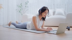 Young Asian woman lying on the floor on yoga mat in front of laptop and training at home. Beautiful smiling woman doing stretching exercise while communicates with a trainer online before workout - Powered by Shutterstock - Get 15% off with code: PIKWIZARD15