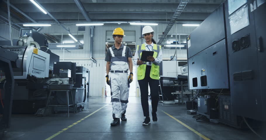 Two Male and a Female Factory Employees Walk Through a High Tech Manufacturing Facility, Discussing Operations and Production Machinery. Diverse Employees Wear Safety Gear and Carry a Tablet Computer