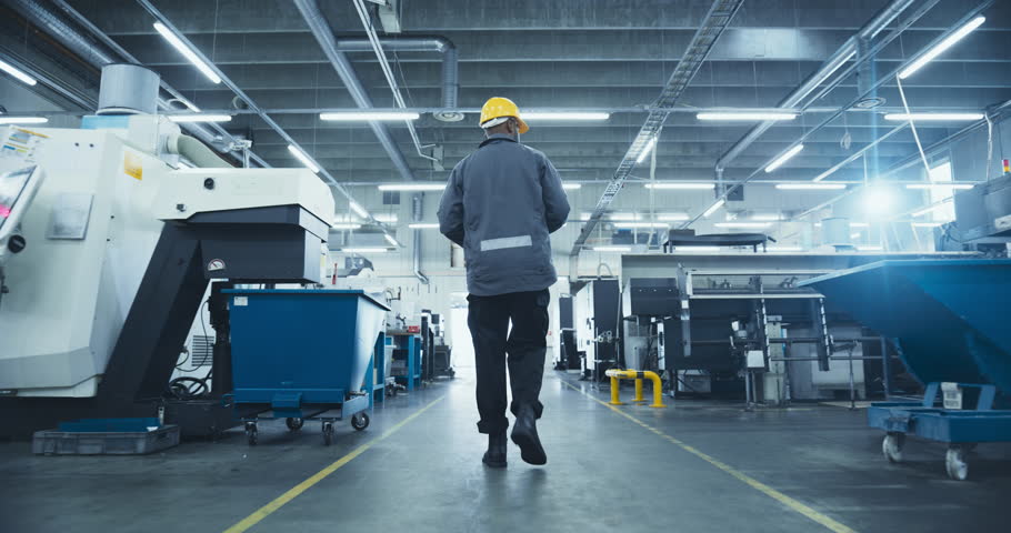 Black Male Worker in a Yellow Hard Hat Walking Through a Modern, Well-lit Factory, Inspecting Advanced Machinery, Checking Equipment, Working in Production Industry. Footage From the Back