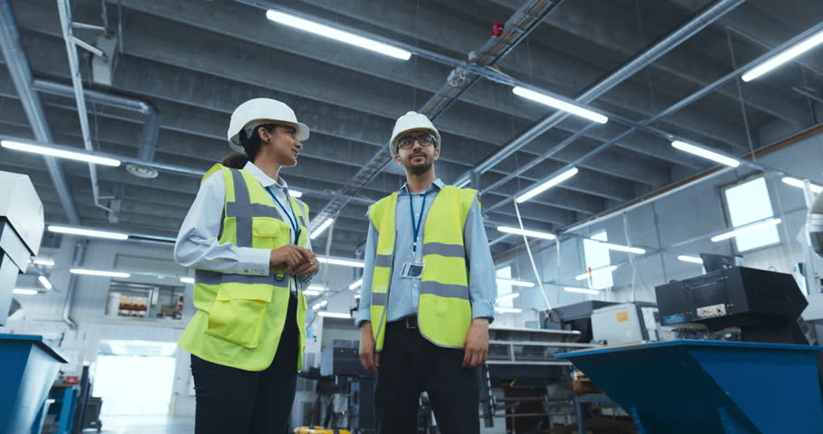 Female Industrial Engineer Meeting with a Production Technologist in a Modern Factory. Colleagues Having a Conversation at Work Amidst Advanced Machinery and Heavy Duty Equipment. Zoom Out Footage