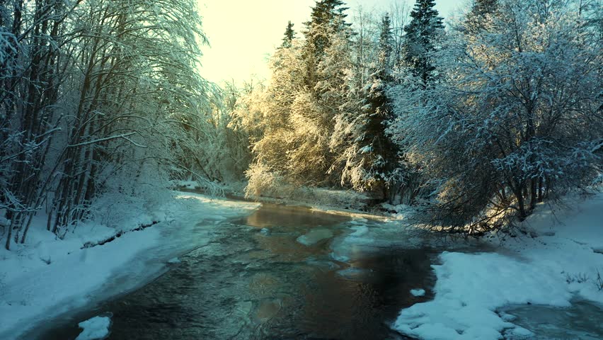 Golden hour sunset glow spreads between trees across flowing river with icy banks and snow