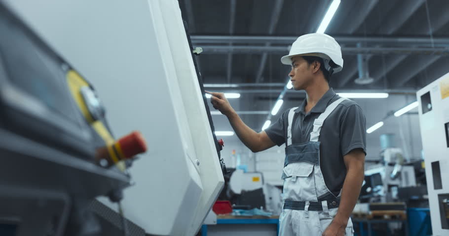 Asian Male Engineer in a White Hard Hat Operating a CNC Milling Machine in a Modern Factory. Young Technician Inserting Parameters into the Computer, Automating Production Line