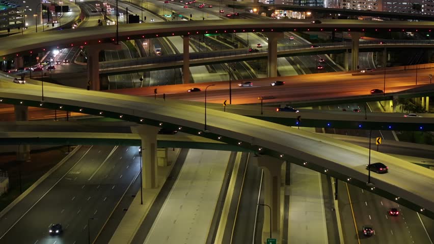 Nighttime aerial drone footage of a multi-level freeway interchange in downtown American city showcasing the intricate network of overpasses and lit-up roads.