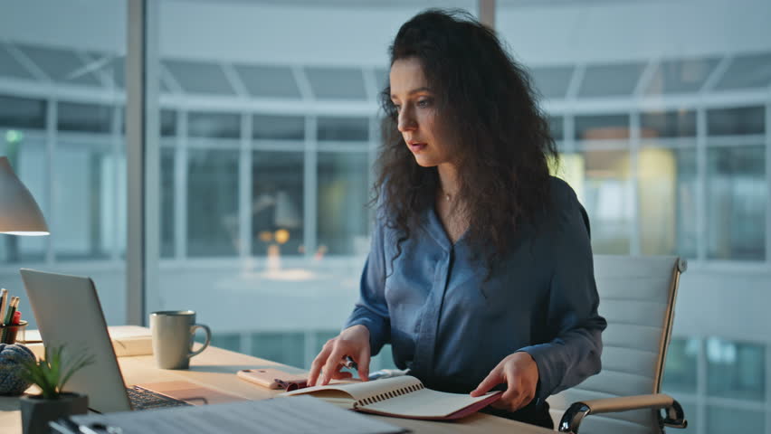 Focused woman working desk in evening closeup. Curly busy employee using laptop in office. Professional manager concentrating reviewing documents. Brunette businesswoman managing tasks at night.