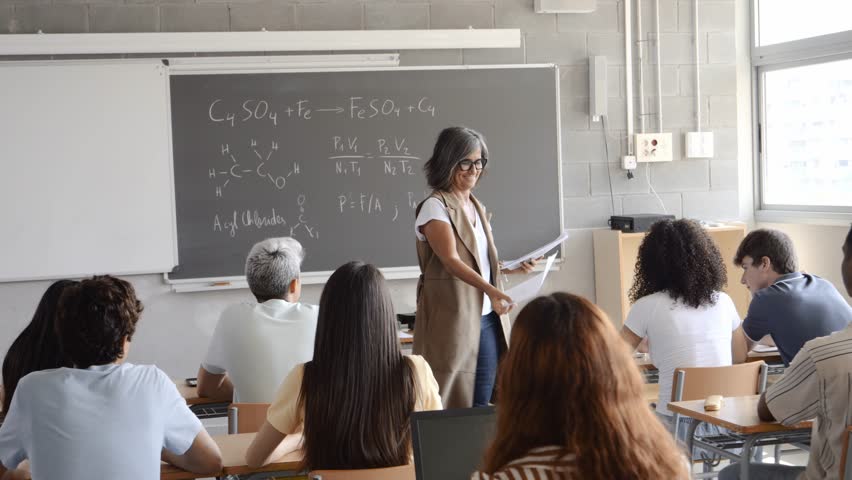 Female teacher handing out exams to a class of diverse teenagers. High school, university, academic exams