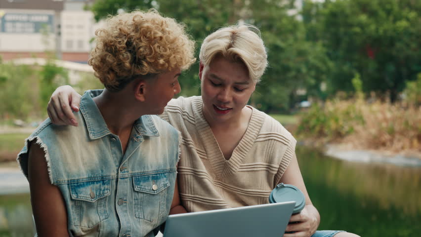 Hapy Asian LGBTQ friends using a laptop and enjoying coffee together in a park. Celebrating pride and connectivity