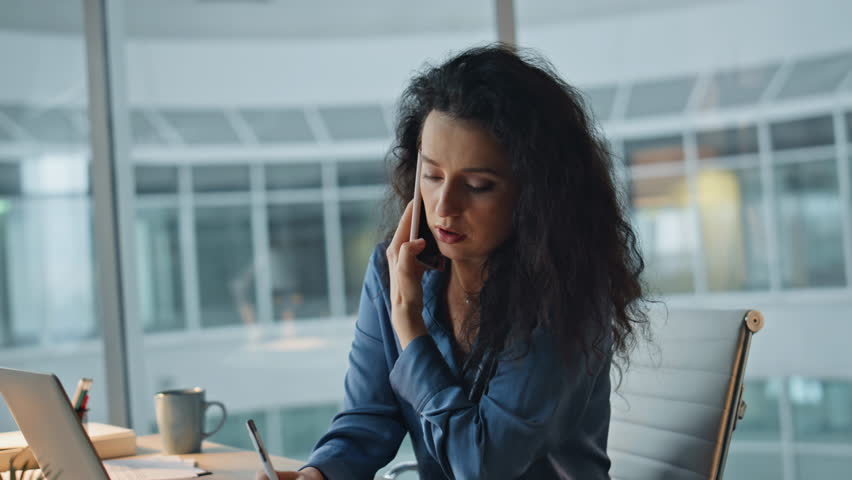 Corporate manager speaking smartphone in evening. Closeup busy executive discuss solving problem in office. Confident professional woman looking at phone taking notes at desk. Productivity concept