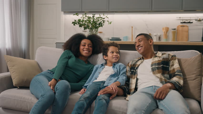 Relaxed tired African American family man dad male mother female woman little child boy son kid falling jumping on comfortable sofa couch rest celebrating own home rented relocation moving living room