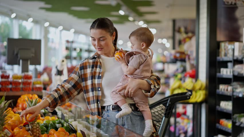 Happy brunette woman in a plaid shirt holds a small child in her arms and shows him orange fruits in a supermarket department