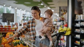 Happy brunette woman in a plaid shirt holds a small child in her arms and shows him orange fruits in a supermarket department - Powered by Shutterstock - Get 15% off with code: PIKWIZARD15