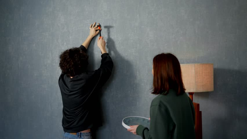 Confident brunette guy hammers a nail into the wall and hangs a clock with the support of his girlfriend in a modern apartment