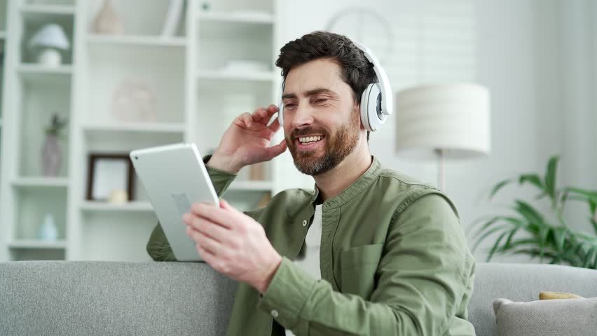 Joyful handsome man wearing wireless headphones listening music using digital tablet sitting on sofa in living room at home. Happy bearded male turns on a playlist and enjoyin favorite song. Close up