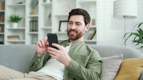 Happy handsome man using mobile phone lying on cozy sofa in living room at home. Smiling male browses social networks, chats online, watches something interesting, writes reads messages on smartphone - Powered by Shutterstock - Get 15% off with code: PIKWIZARD15